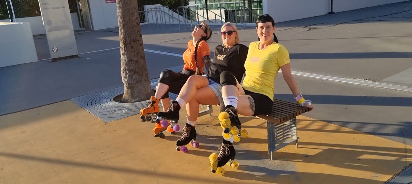 Skate Pink rollerskating tees to suit your whole skate squad.  Three skater girls in orange, black and yellow Skate P shirts, sat on a park bench, legs crossed with roller skates on.  Their tees match their skates.